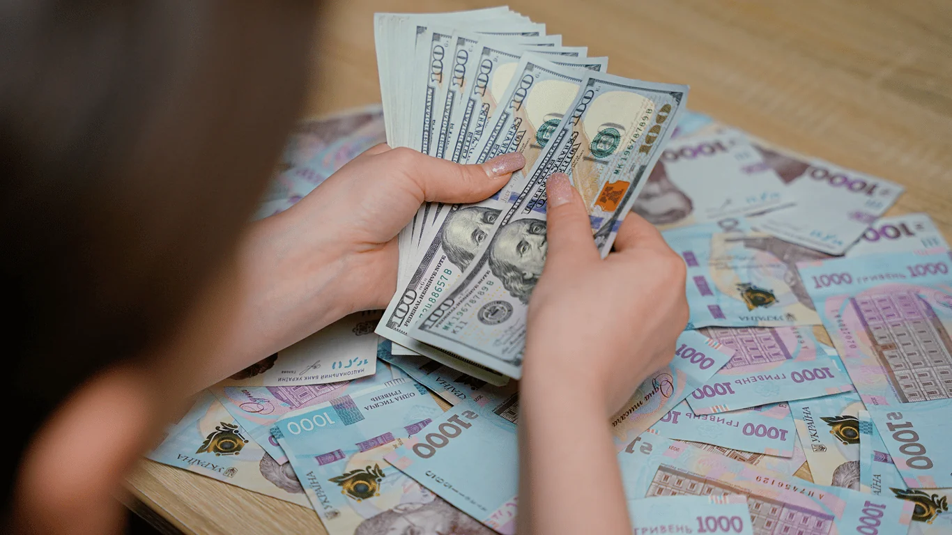 Руки перераховують стодоларові купюри на тлі українських гривень