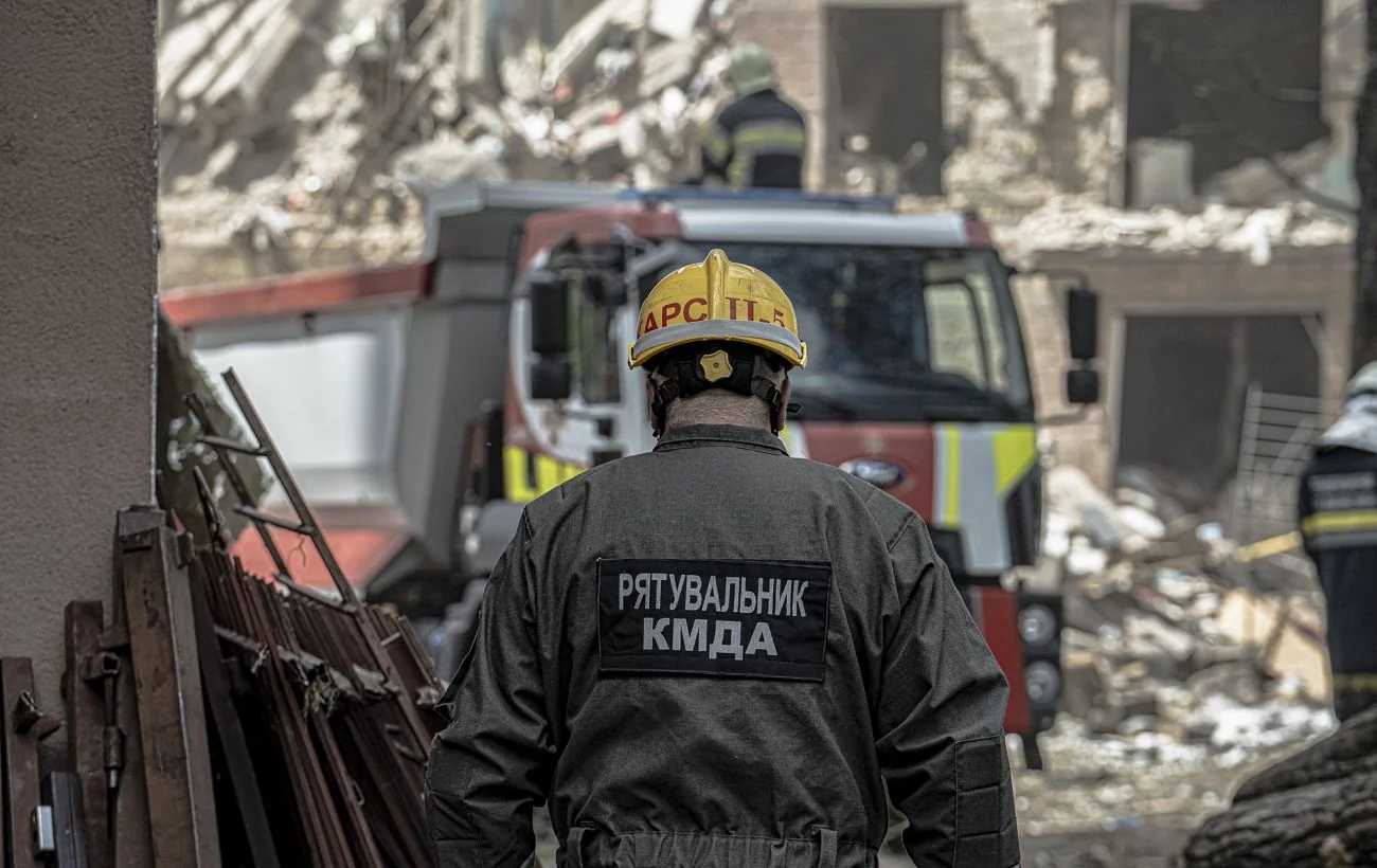 Рятувальник КМДА в жовтому шоломі біля пожежної машини на тлі зруйнованих будинків у Києві