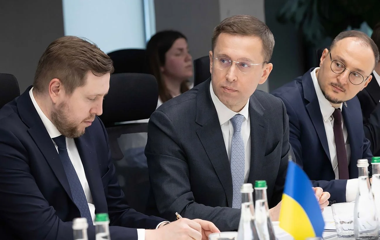 Троє чоловіків у костюмах за столом наради з українським прапором